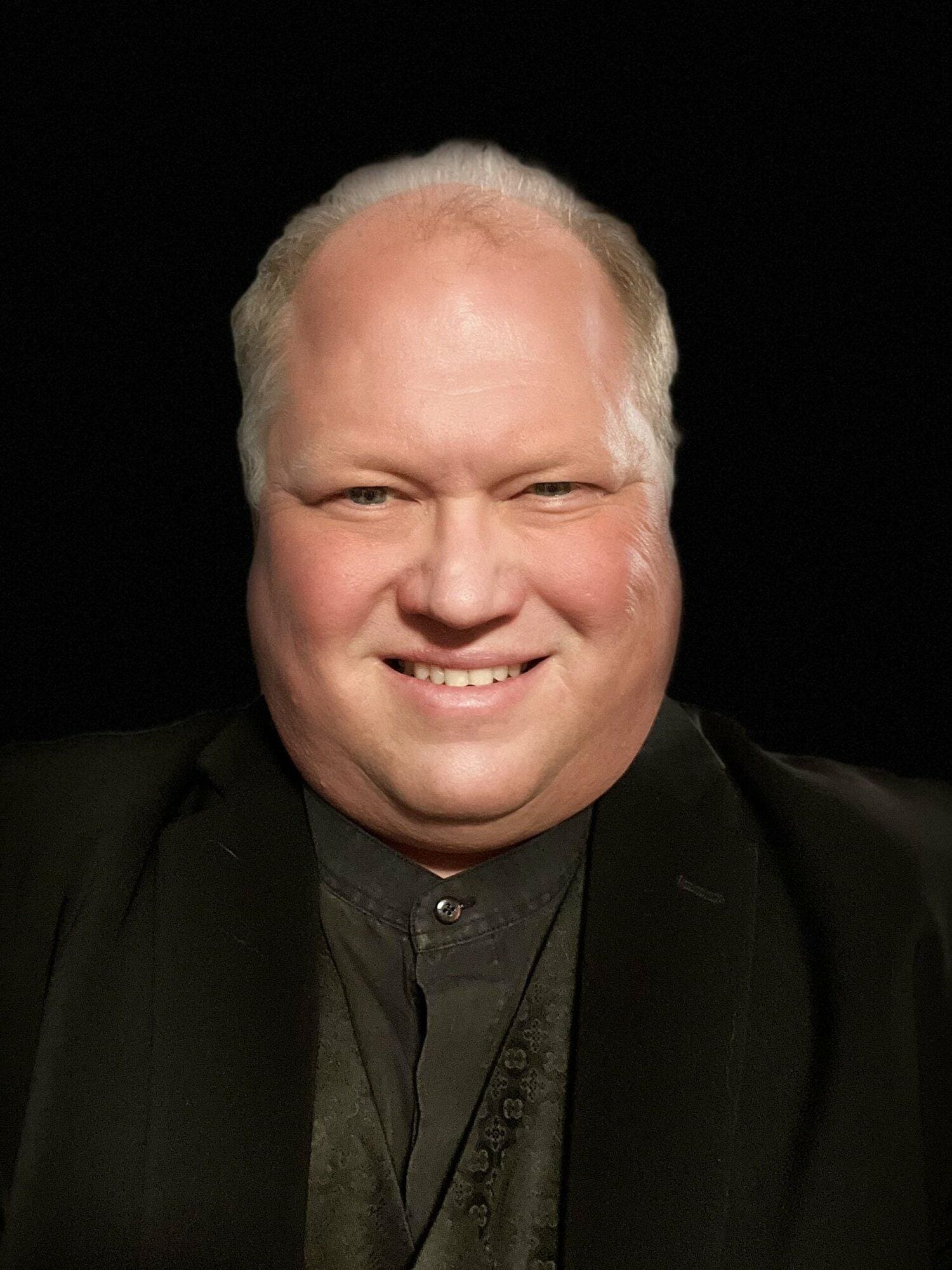 A person with light skin tone, short light-colored hair, and a black suit smiles at the camera against a black background.
