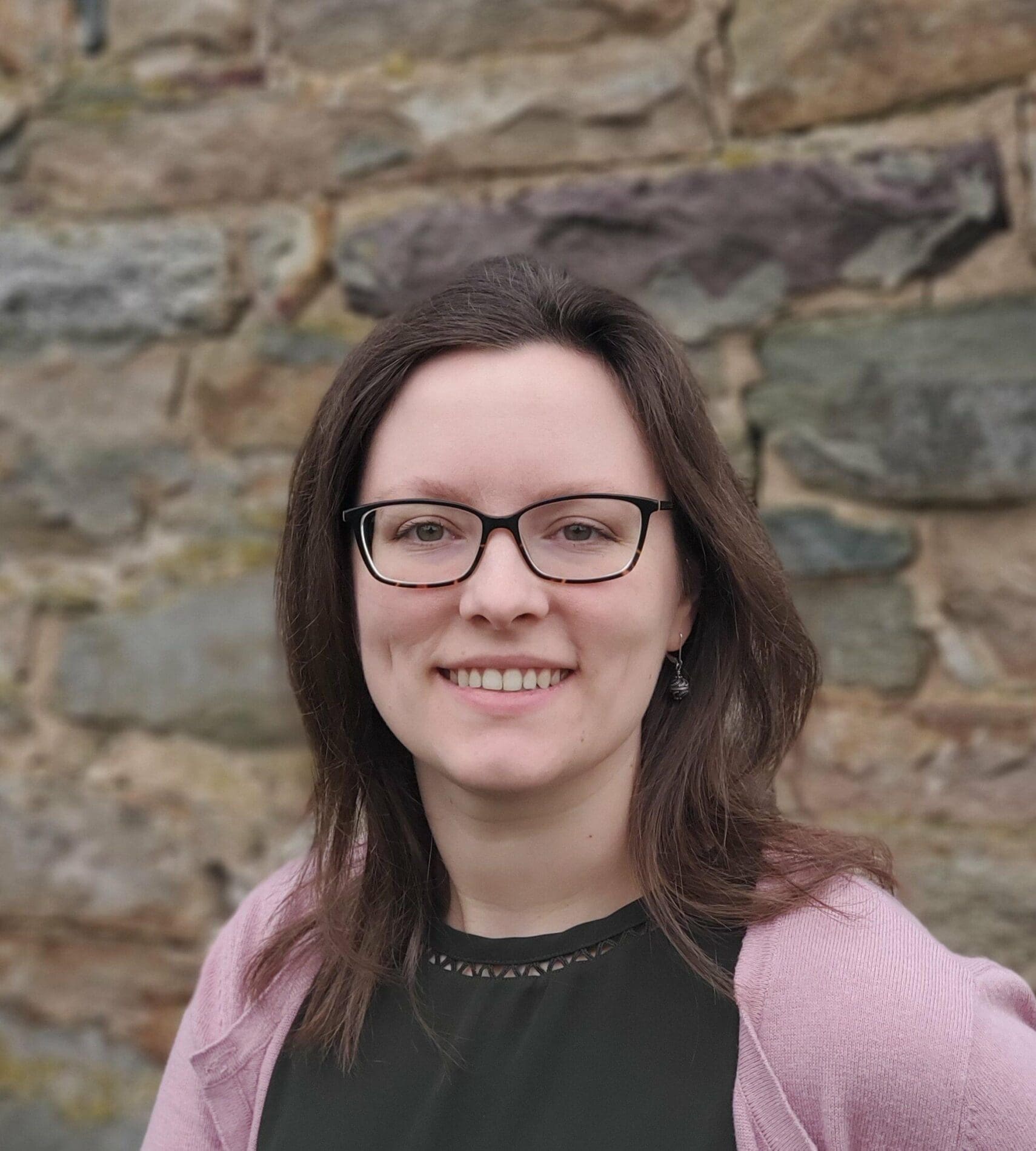A person with glasses and shoulder-length hair is smiling in front of a stone wall. They are wearing a black top and a light pink cardigan.