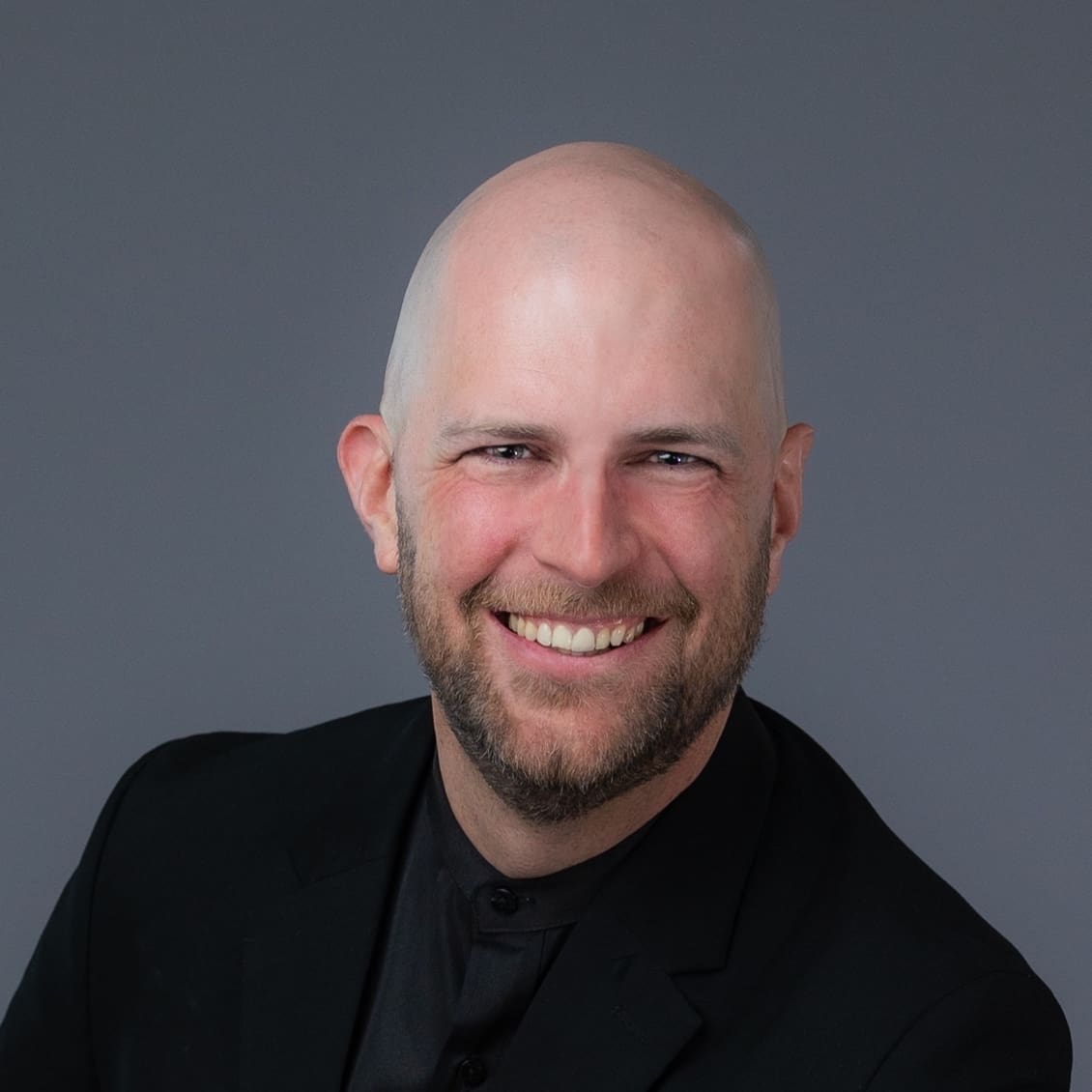 A bald man with a beard is smiling, wearing a black suit against a gray background.