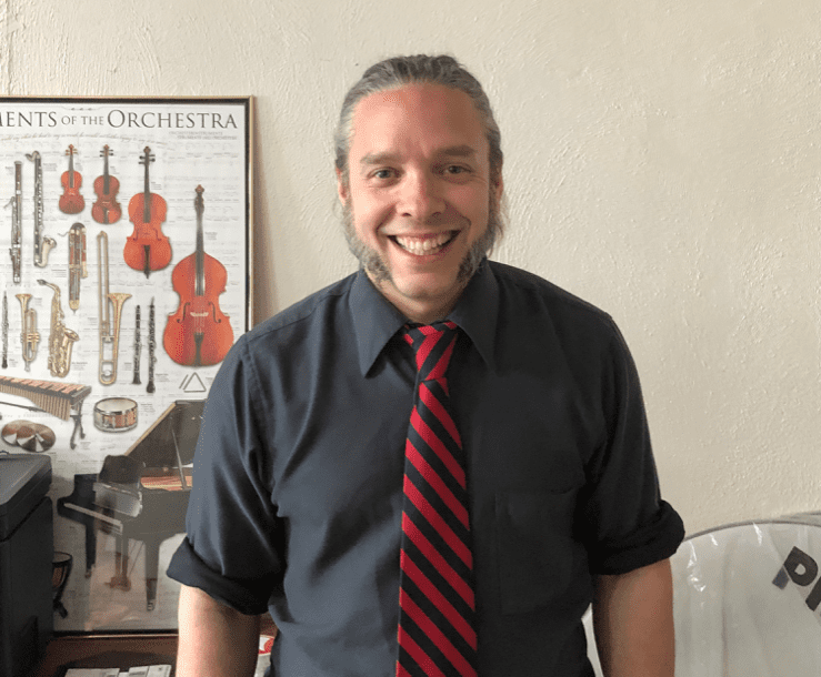 Smiling man with a gray shirt and red-striped tie stands in front of a wall poster featuring orchestral instruments.
